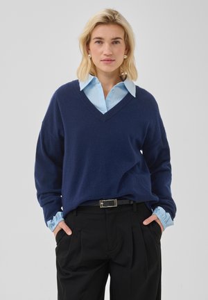 Frau mit schulterlangem blondem Haar trägt einen marineblauen V-Ausschnitt-Pullover über einem blauen Hemd mit Kragen und schwarze Hose, Hände in den Taschen, neutraler Hintergrund.