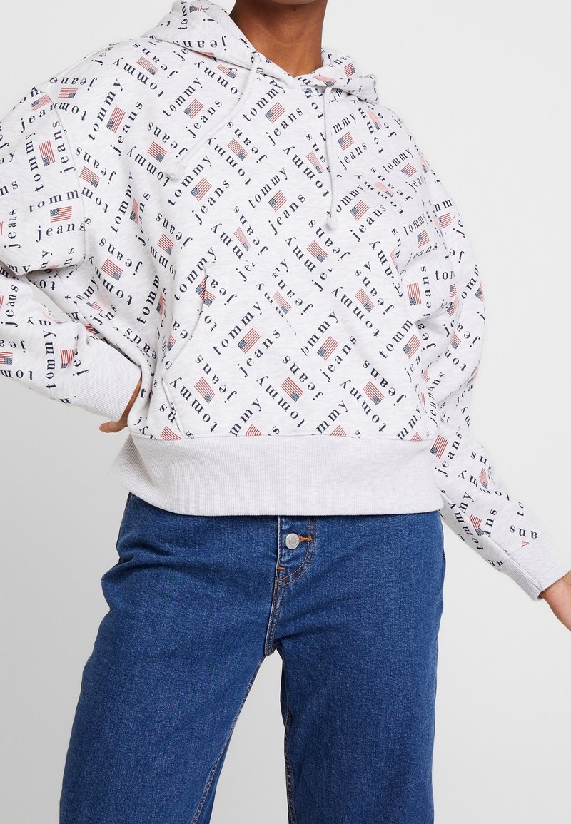 Personne portant un sweat à capuche blanc avec le texte répété "Tommy Jeans" et des petits motifs du drapeau américain, associé à un jean en denim bleu.
