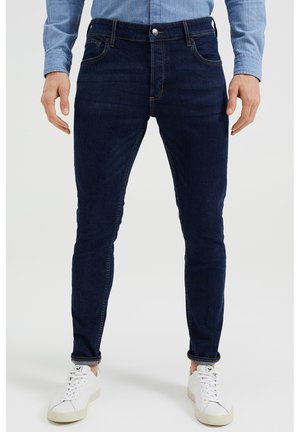 Man draagt donkerblauwe slim-fit jeans met opgerolde pijpen, een lichtblauw shirt met lange mouwen en witte sneakers, staand tegen een effen achtergrond.