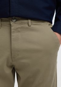 Person iført beige bukser med knap foran og lommer, hånd hvilende i højre lomme, kombineret med en mørkeblå skjorte.