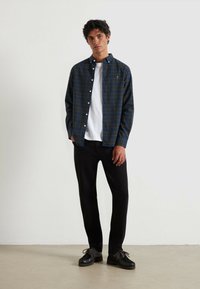 Jonge man staat tegen een witte muur, gekleed in een marineblauw en groen geruit overhemd over een wit T-shirt, zwarte broek en zwarte schoenen.
