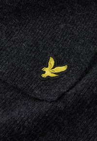Tessuto di maglia nera con un distintivo logo di aquila ricamato giallo su una superficie strutturata, con un dettaglio della tasca visibile.