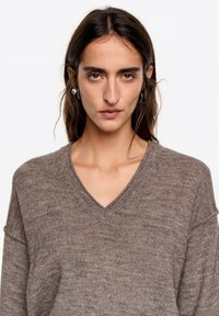 Luźny sweter z dekoltem w serek w stonowanym brązowym odcieniu, wykonany z miękkiego, teksturowanego dzianego materiału. Rękawy są lekko opuszczone.