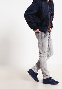 Veste bomber bleu marine, chemise vert olive, jean gris clair et baskets bleu marine avec unité d'air visible, présentant un design élégant et décontracté.