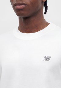 Közeli kép egy személyről, aki fehér New Balance kereknyakú pólót visel, amelyen az NB logó hímezve van a mellkason.
