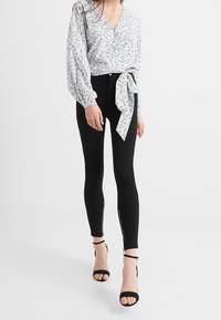 Blouse blanche avec motif floral, décolleté en V, et lien à la taille. Jean skinny noir avec zips sur les côtés. Talons aiguilles noirs à lanières.
