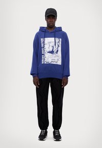 Μπλε φούτερ με γραφικά, κουκούλα με κορδόνι και μακριά μανίκια, συνδυασμένο με μαύρο παντελόνι με διακριτικό σχέδιο και μαύρα αθλητικά παπούτσια.