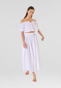 Fehér, vállnélküli crop top fodros kialakítással, hosszú, légies fehér szoknyával párosítva, amely rugalmas derékpánttal rendelkezik.