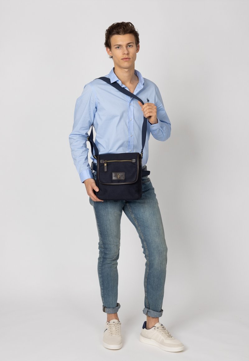 Borsa messenger navy con patta anteriore, chiusura con zip e una placca logo metallica. Realizzata in tessuto resistente, presenta una lunga tracolla regolabile.