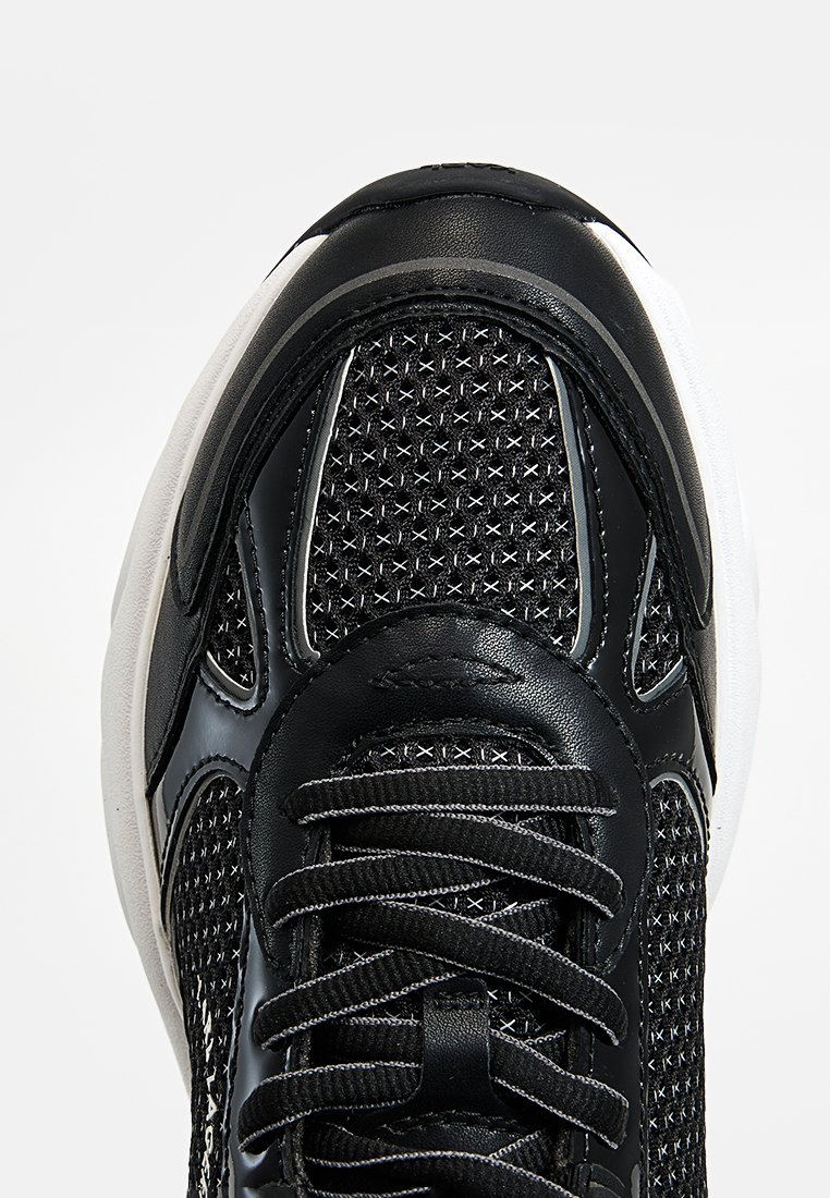 Zapatilla deportiva negra con una parte superior de malla que presenta un patrón de diamante texturizado, acentos de cuero suave y una suela blanca contrastante.
