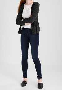 Svart kofta över en vit topp, tillsammans med mörka tajta jeans och svarta slip-on skor. Släta texturer och figursydd design.