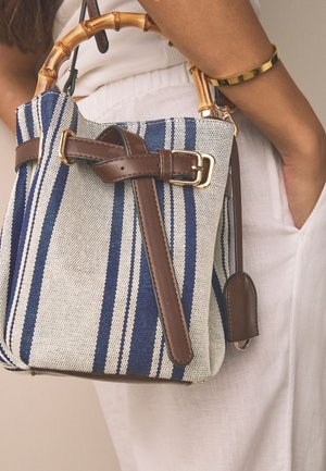 Vrouw houdt een blauw-wit gestreepte stoffen tas met bamboehendels en een bruine leren band vast, draagt witte broek en een gouden armband.