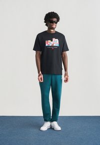 Μαύρο βαμβακερό T-shirt με γραφικό σχέδιο, σε συνδυασμό με τιρκουάζ φόρμα. Το μοντέλο φοράει γυαλιά ηλίου και λευκά αθλητικά παπούτσια. Το σκηνικό είναι ουδέτερο.