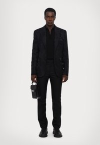 Black blazer with floral pattern, black polo, tailored trousers, and black shoes. Holding a small black handbag with silver accents.