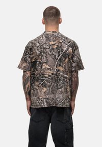 Camo-patterned short-sleeve t-shirt featuring shades of brown, gray, and green, with a loose fit and soft fabric texture. Circular neckline.