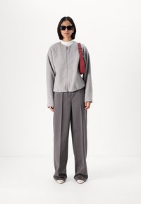 Pelēka pūkaina jaka ar rāvējslēdzēju virs baltā turtlenecka, saskaņota ar augsto jostasvietu pelēkiem biksēm un bēšiem šauriem apaviem. Melni saulesbrilles un sarkana soma piešķir akcentu.
