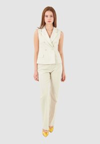 Blazer senza maniche beige chiaro con doppi bottoni, abbinato a pantaloni attillati coordinati e décolleté gialli a punta. Strisce verticali sottili visibili.