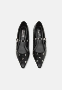 Chaussures en cuir vegan noir avec un bout pointu, ornées d'œillets en argent et d'une sangle décorative au milieu du pied. Texture lisse.