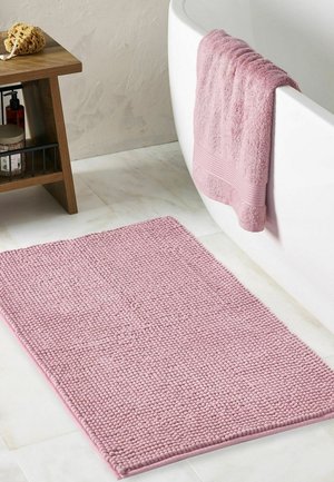 Hellrosa strukturierte Badematte auf weiß gefliestem Boden neben einer weißen Badewanne mit passendem rosa Handtuch, das über den Rand hängt. Holzregal mit Badeartikeln in der Nähe.
