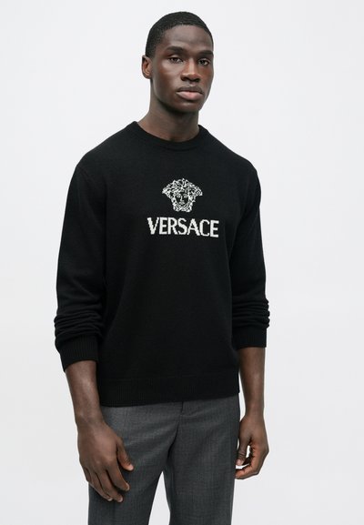 Černý svetr z pleteného materiálu s bílým vyšívaným logem Versace a motivem Medúzy na předn�í straně. Modelka má na sobě šedé kalhoty.