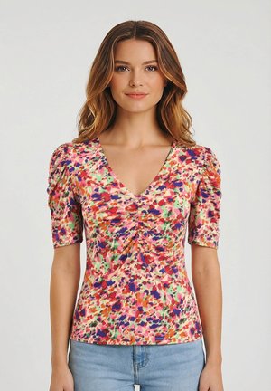 Femme portant un chemisier multicolore à manches courtes avec un col en V et un devant froncé, associé à un jean bleu clair, debout devant un fond uni.