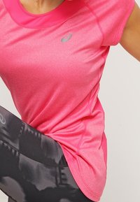 Pink short-sleeve athletic shirt with a smooth texture, featuring a contrasting neckline and a reflective logo. Worn with black patterned leggings.