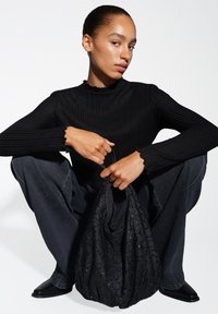 Zwarte geribbelde turtleneck gecombineerd met losse donkere jeans. Het model houdt een zwarte pailletten tas vast met een zachte textuur en geplooid ontwerp.