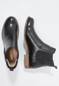 Schwarze Leder-Stiefeletten mit leichtem Glanz, elastischen Seiteneinsätzen und einem niedrigen, gestapelten Holzabsatz, ausgestattet mit einer Schlaufe zum einfachen An- und Ausziehen.