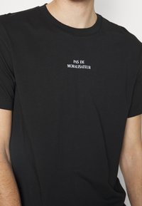 Czarna bawełniana koszulka z białym napisem na piersi, który brzmi "PAS DE MORALISATEUR." Klasyczny okrągły dekolt i krótkie rękawy.