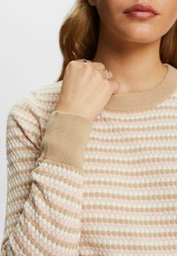 Lekko teksturowany sweter z wzorem pastelowych okręgów na beżowym tle. Posiada ściągacze przy mankietach i dekolcie w dopasowanym kolorze.