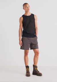 Débardeur noir côtelé, shorts gris et bottes en cuir marron à lacets avec des accents jaunes. Le modèle se tient avec une posture détendue contre un fond uni.
