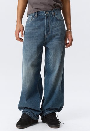 Persona que lleva puestos unos vaqueros azules desgastados y holgados con zapatillas negras, de pie contra un fondo claro y sencillo.