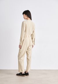 Combinaison beige avec une taille ceinturée, des poignets boutonnés et une coupe décontractée. Portée avec des chaussures noires comportant de subtils accents métalliques.