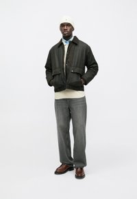 Mann in braunem Jackett, hellblauem Hemd, cremefarbenem Pullover, grauen Jeans, weißer Strickmütze und braunen Lederschuhen, der mit den Händen in den Taschen dasteht.