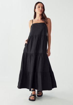 Abito maxi nero con spalline sottili, design a strati e texture morbida. Dotato di tasche e una silhouette fluida, abbinato a sandali neri.