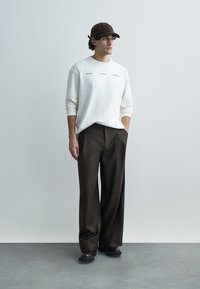 Bela srajca z dolgimi rokavi z napisom, rjave široke hlače in rjava kapa. Model stoji z rokami v žepih na nevtralnem ozadju.