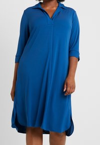 Woman wearing a loose-fitting, knee-length blue dress with three-quarter sleeves and a collar, standing against a plain background.