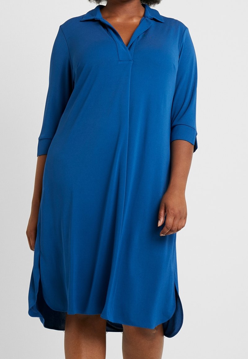 Woman wearing a loose-fitting, knee-length blue dress with three-quarter sleeves and a collar, standing against a plain background.