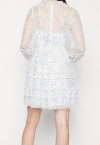 Robe blanche avec des manches longues transparentes, des accents volantés et une jupe superposée. Présente un motif floral et un col haut. Vue arrière montrée.