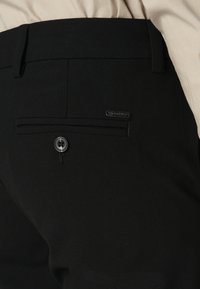 Nahaufnahme von schwarzen, maßgeschneiderten Hosen mit einer Gesäßtasche, die einen Knopf und ein metallisches Markenlabel aufweist, getragen mit einem beigen Hemd.