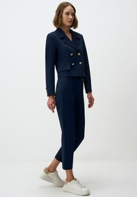 Marineblauwe dubbelgebreide blazer met gouden knopen, gecombineerd met nauwsluitende marineblauwe broek en witte sneakers. Glad stof met een op maat gemaakte uitstraling.