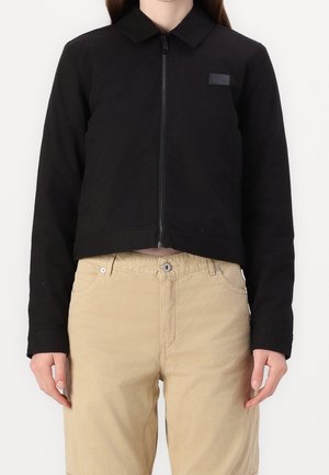 Kvinde iført en sort lynlåsjakke med krave og beige bukser, stående mod en ensfarvet hvid baggrund.