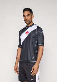 Kratka športna majica s kratkimi rokavi v temno sivi barvi z belim diagonalnim panelom in rdečim križem. Izdelana iz gladkega, lahkega materiala.