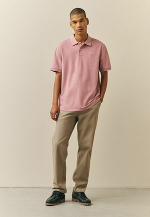 Bărbat tânăr stând sprijinit de peretele bej, purtând un tricou polo roz deschis, pantaloni bej și pantofi negri cu șiret, mâinile relaxate pe lângă corp.