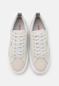 Ett par ljusbeiga Hugo Boss sneakers med vita snören, mesh tyg, läderpaneler och vita sulor, sedd från ovan.