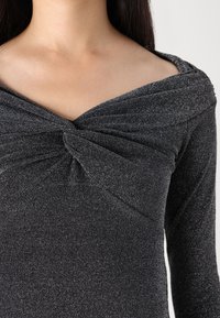 Black knitted top with a deep V-neck, featuring a twisted front detail, long sleeves, and a textured fabric. Soft, metallic accents throughout.