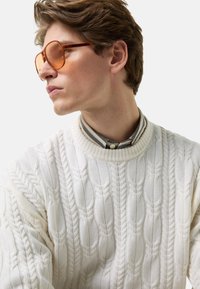 Cremefarbener, kabelgestrickter Pullover mit einem gerippten Ausschnitt, getragen über einem grau gestreiften Hemd. Übergroße Sonnenbrille mit orangefarbener Tönung.