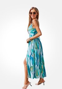 Zomerse jurk met een getailleerde bodice en een vloeiende rok, voorzien van een blauw en groen bladerenpatroon op lichtgewicht stof. Witte hoge hak sandalen.