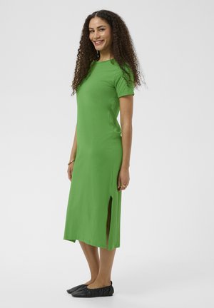 Femme aux cheveux bouclés souriante, portant une robe midi verte à manches courtes avec une fente latérale et des chaussures plates noires, debout de profil.