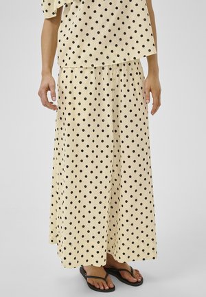 Beiges Rock- und Top-Set mit schwarzen Polka-Dots, getragen mit schwarzen Flip-Flops, gezeigt vom mittleren Oberkörper bis zu den Füßen vor einem einfarbigen Hintergrund.